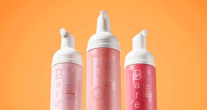 Three pink bottles of Bare by Vogue self-tan foam in Medium, Dark, and Ultra Dark shades, set against a vibrant orange background.
