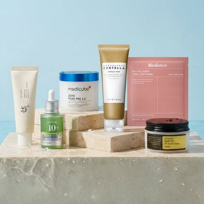 A collection of popular Korean skincare products from brands like Anua, Medicube, SKIN1004, and COSRX, arranged on light stone pedestals against a soft blue background.